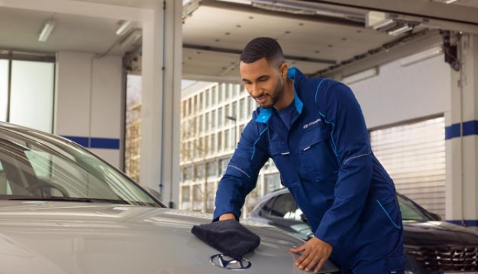 Mechanic in blue overalls polishing the bonnet of a silver Hyundai car in a garage.
