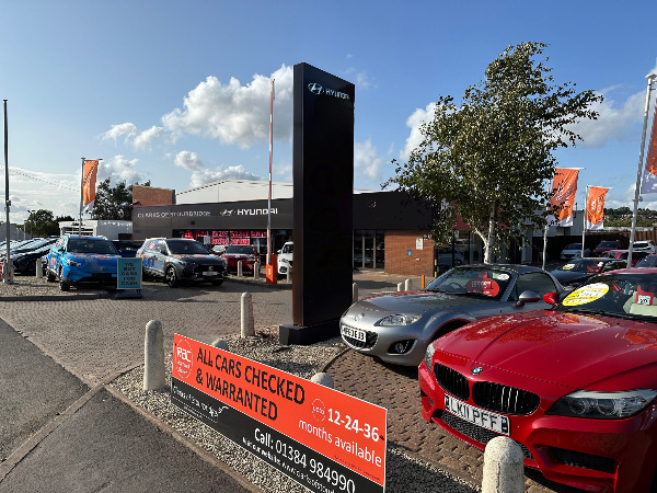 Car dealership with various vehicles on display and an RAC warranty sign in the foreground.