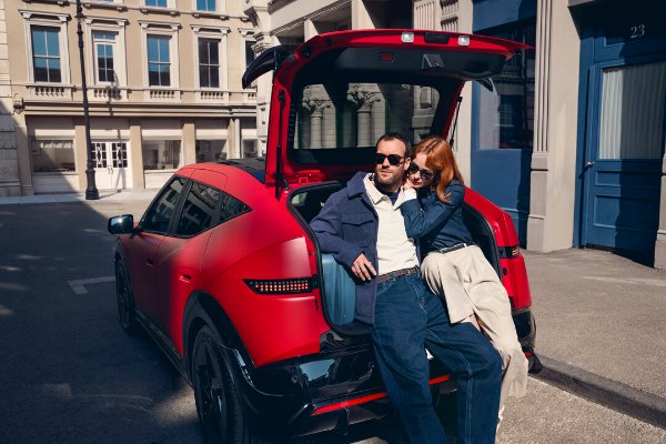 Couple sitting in the open boot of a red car on a city street.